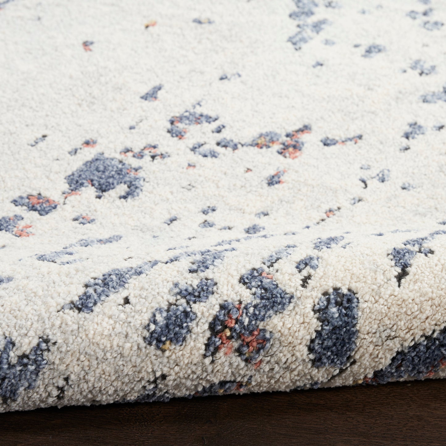 Cream rug with blue-gray & rust blotches; plush textured pile, rolled edge on wooden floor.