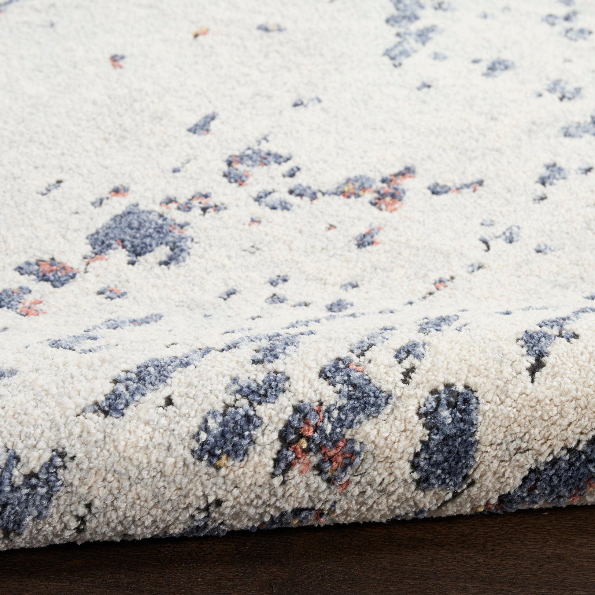 Cream rug with blue-gray & rust blotches; plush textured pile, rolled edge on wooden floor.