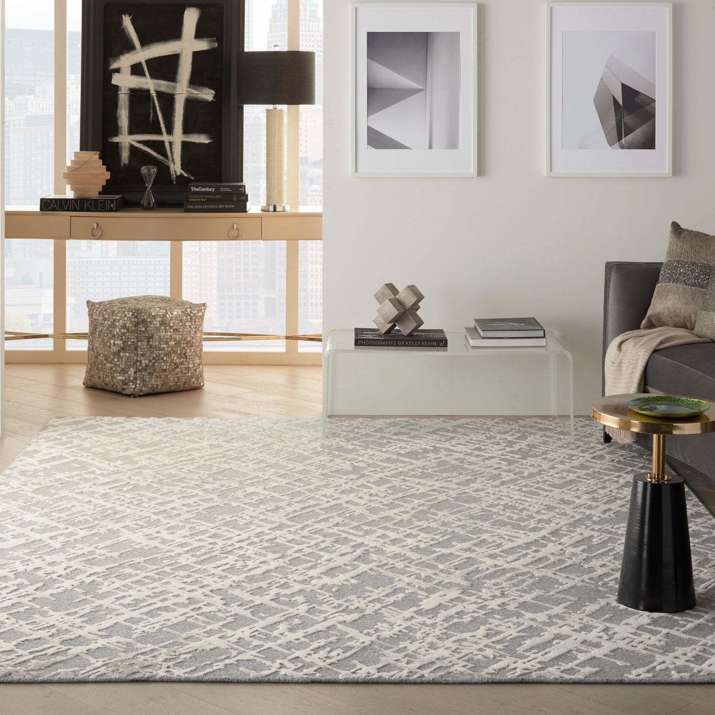 Modern living room with a gray geometric rug, glass coffee table, gray sofa, white-framed abstract art, sculptural decor, city view.