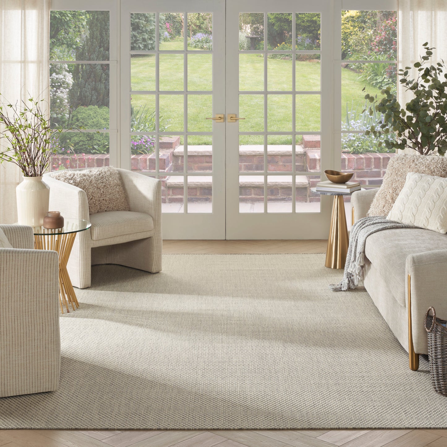 Bright living room with cream sofas, wicker chairs, beige woven rug, glass coffee table, gold accent side tables, potted plants, French doors to garden.