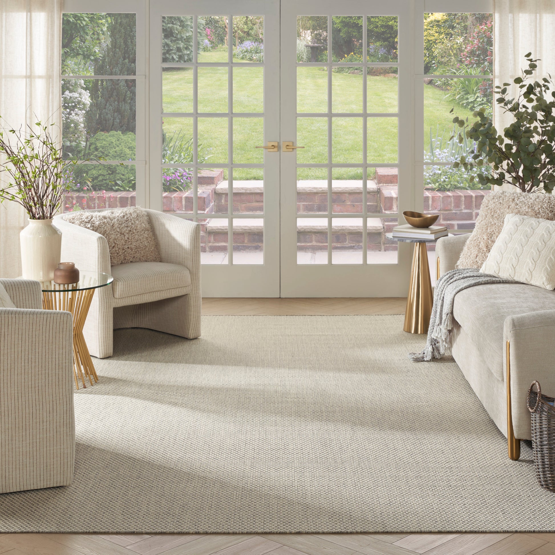 Bright living room with cream sofas, wicker chairs, beige woven rug, glass coffee table, gold accent side tables, potted plants, French doors to garden.
