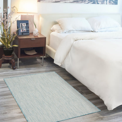 Light blue patterned rug on the floor in a bedroom scene with a white bed, thick comforter and two pillows, a dark wood nightstand with a lamp, plant and photo frame.