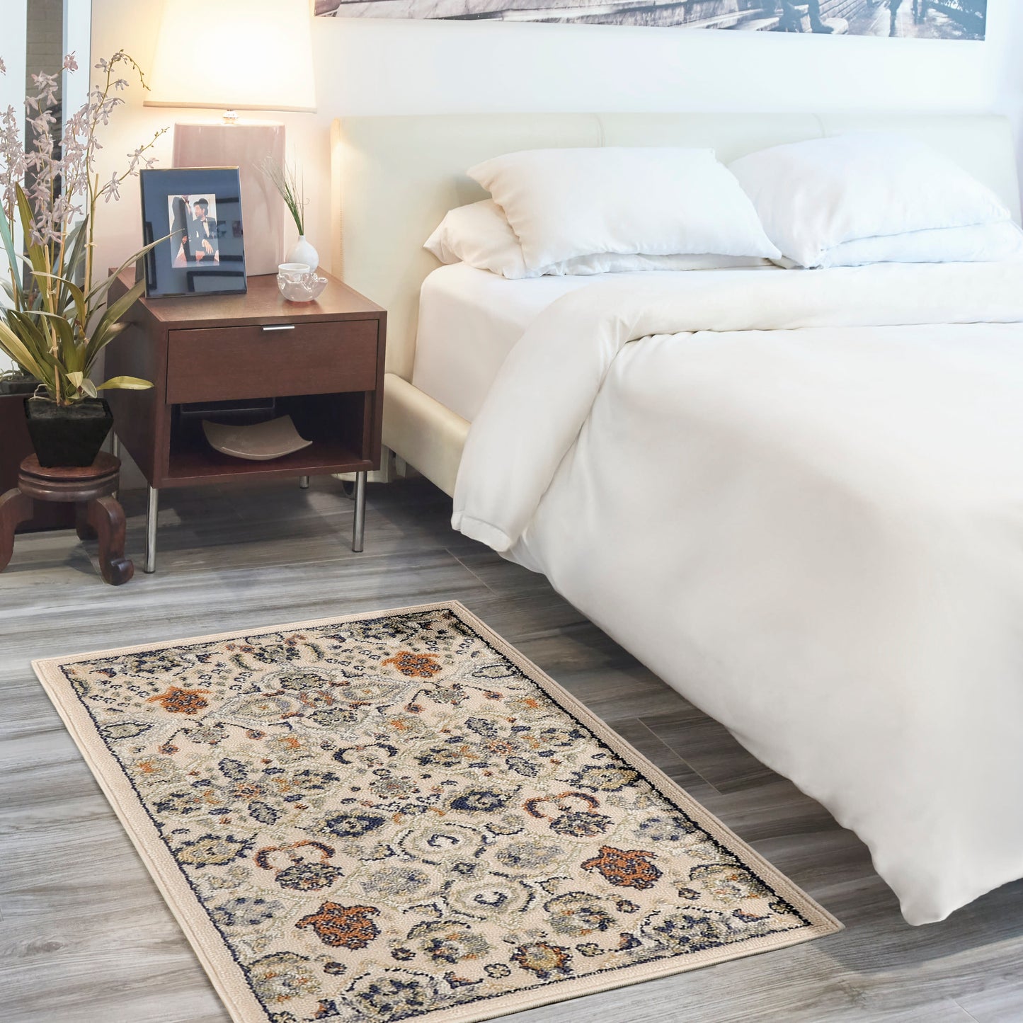 Beige floral area rug (3' x 5') in a bedroom scene with an ivory bed, white duvet and pillows, dark wood nightstand with lamp, plant, and framed photo.