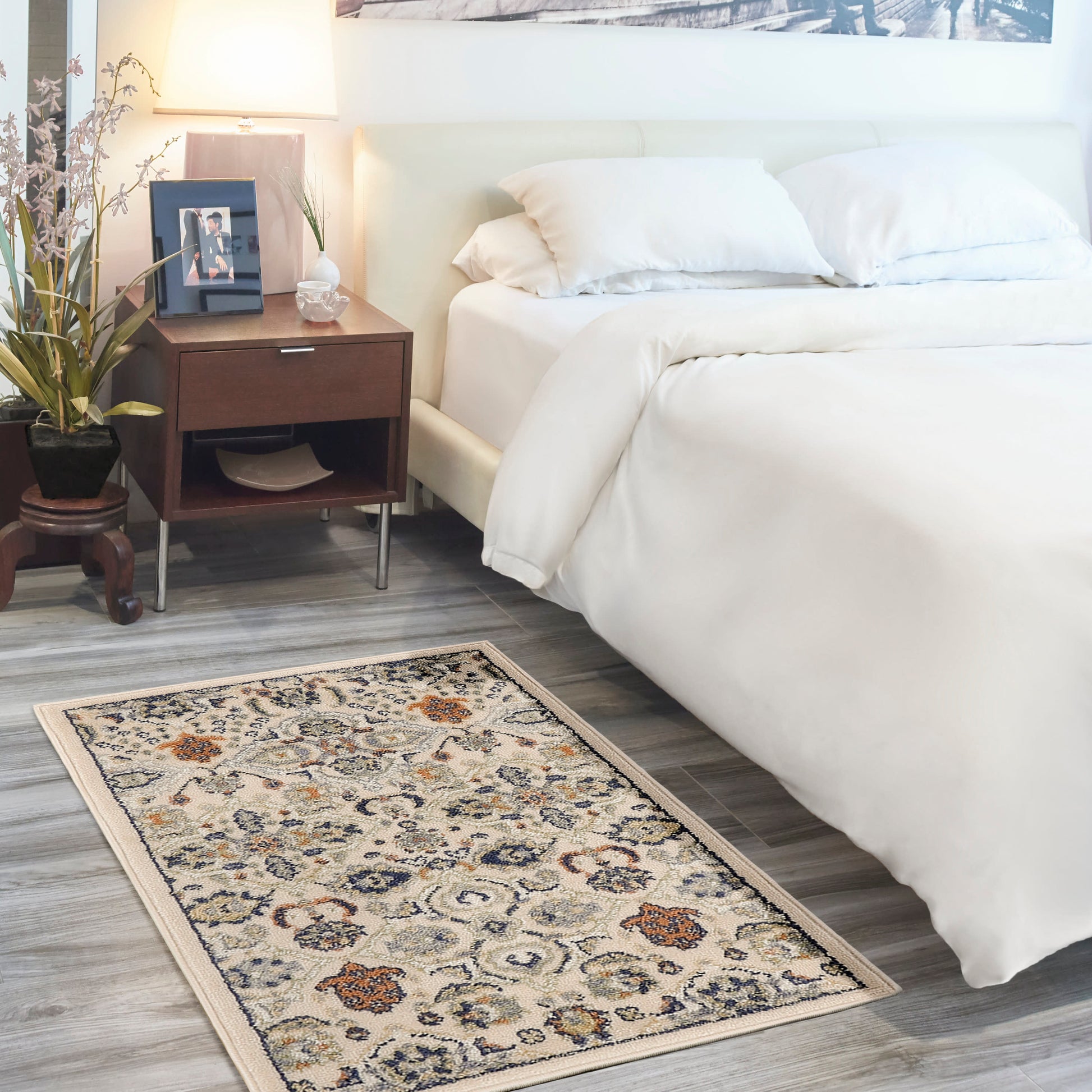 Beige floral area rug (3' x 5') in a bedroom scene with an ivory bed, white duvet and pillows, dark wood nightstand with lamp, plant, and framed photo.