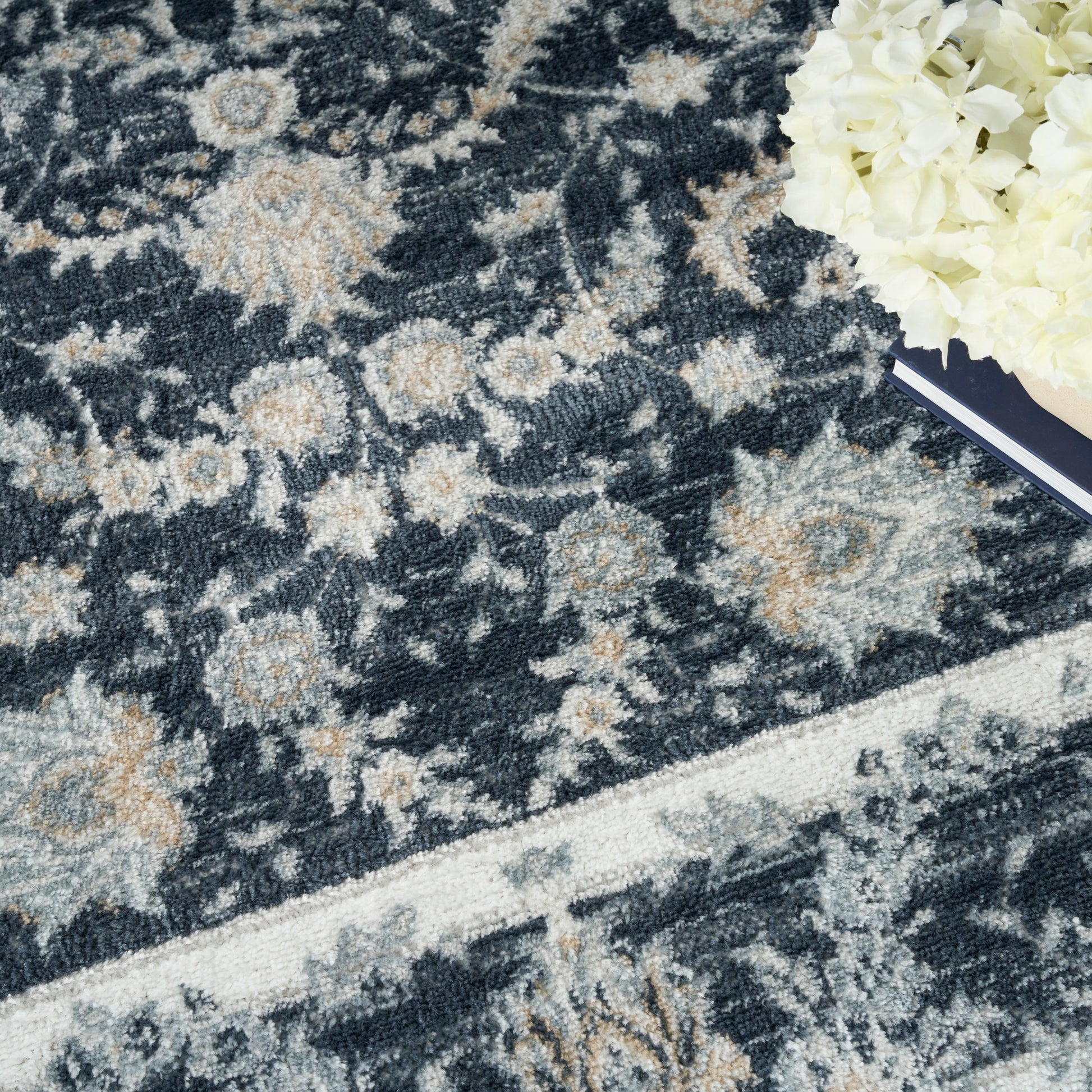 Dark navy oriental rug with beige floral pattern and ivory border; plush pile, white bouquet and a book in the corner.