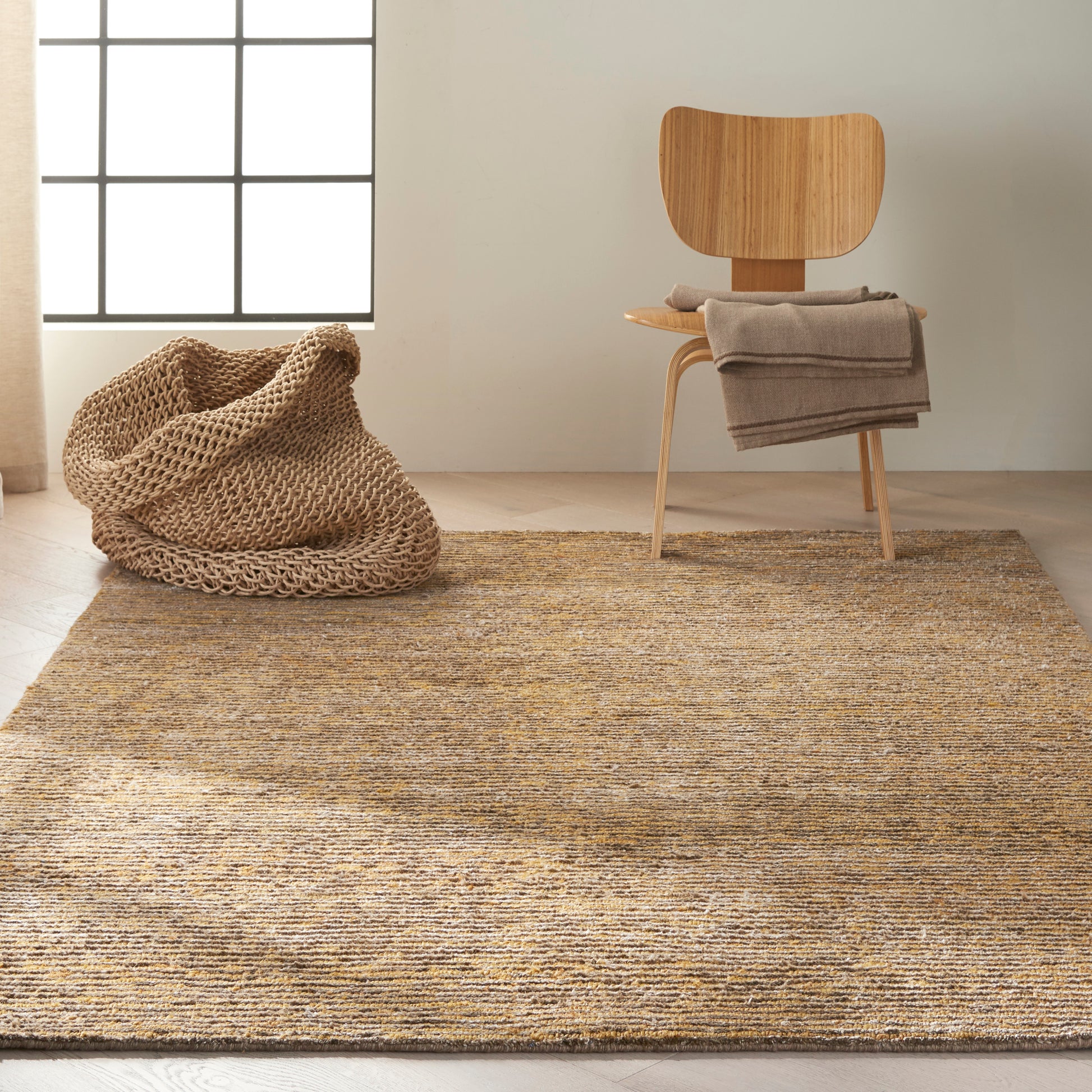 Minimal warm living room: woven jute rug, knitted pouf, wooden chair with gray throw, grid-window light.