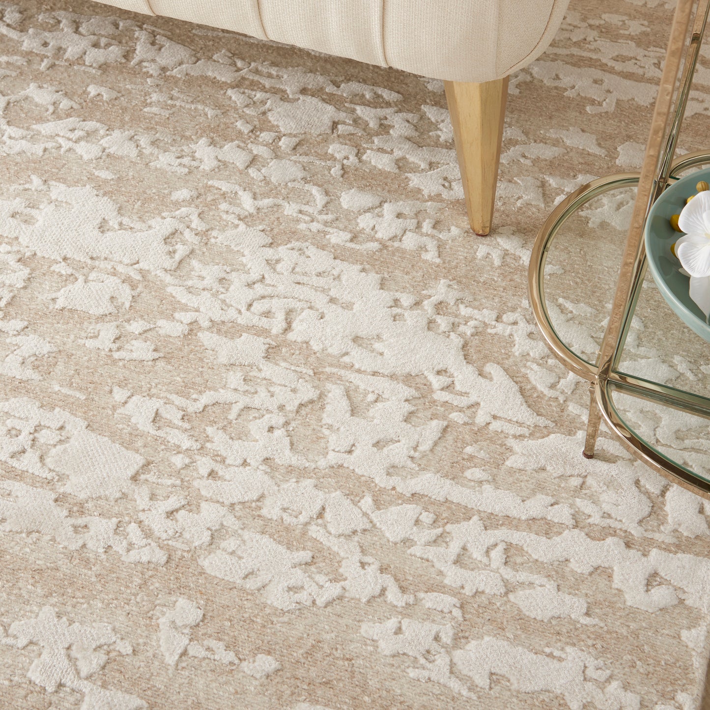 Beige area rug with white abstract shapes, beside a cream sofa leg and a glass round coffee table.