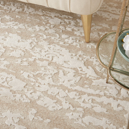 Beige area rug with white abstract shapes, beside a cream sofa leg and a glass round coffee table.