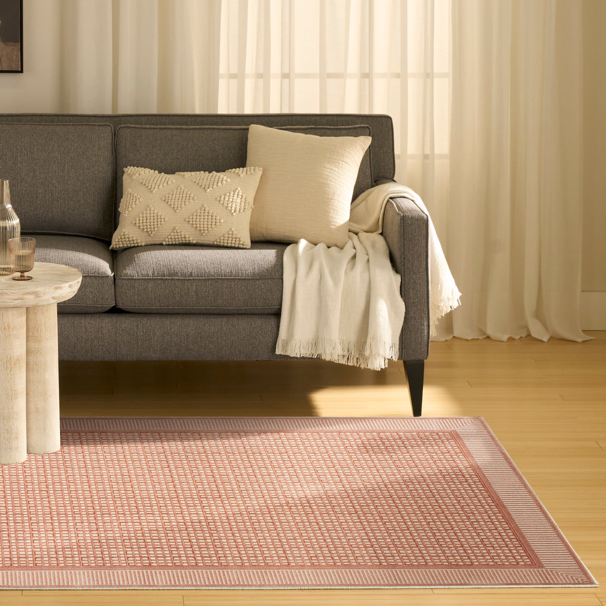 Gray fabric sofa with cream throw and beige cushions, wooden side table, pink-patterned area rug, sheer curtains.
