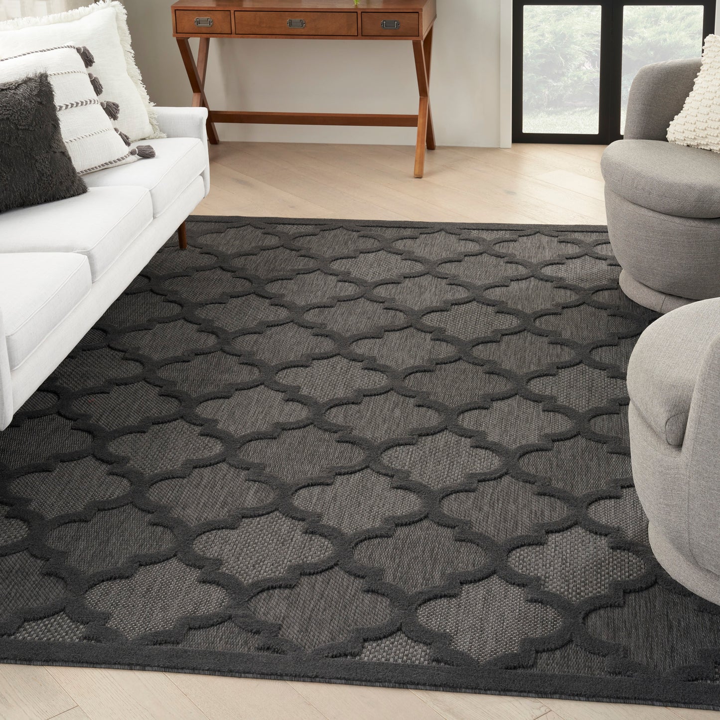 Dark gray geometric trellis rug on light wood floor in a modern living room with white sofa and gray chairs.
