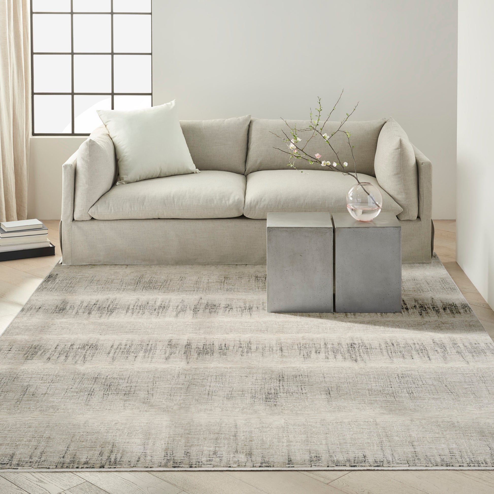 Neutral living room with light gray sofa, pale pillow, two cube gray side tables, vase with branches, textured beige rug.