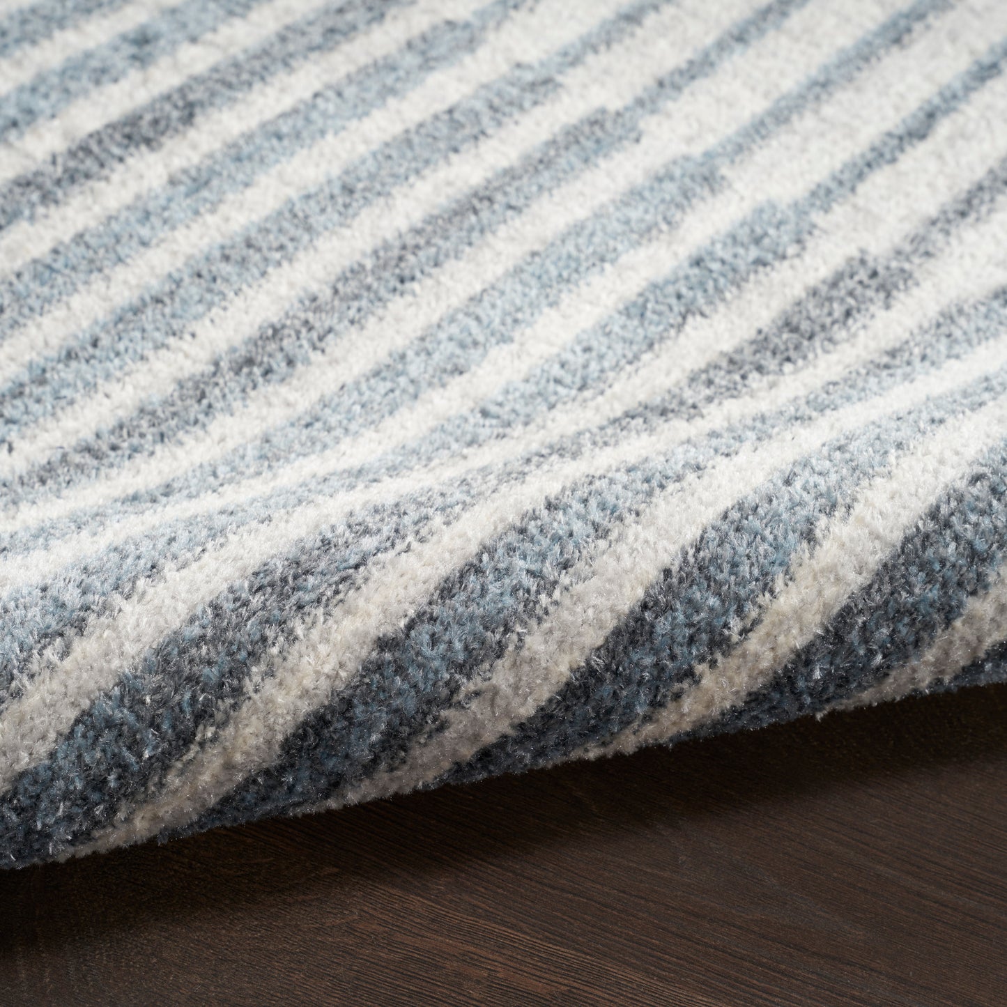 Close-up of blue and gray striped rug with plush pile, edge folded over, on dark wooden floor.