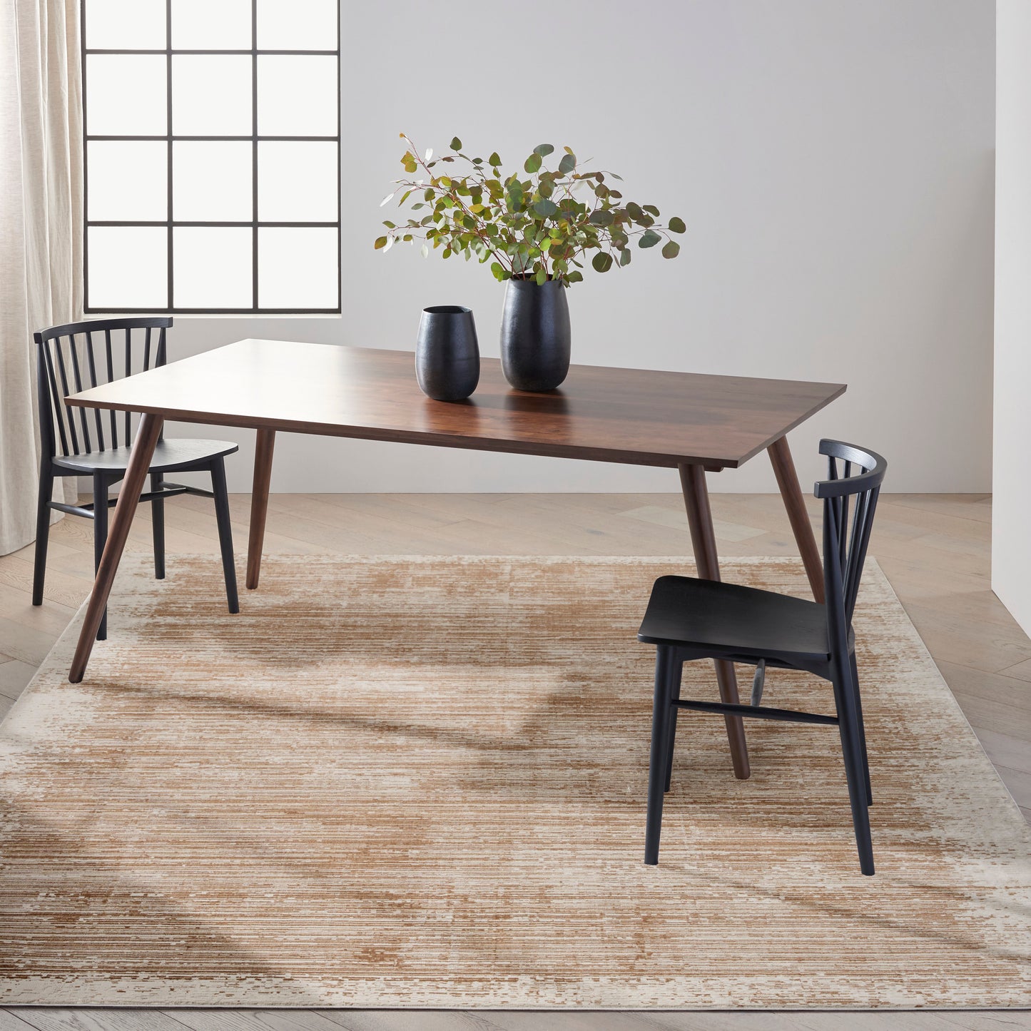 Mid-century walnut rectangular dining table with angled legs, black chairs, and a two-vase greenery centerpiece on a beige rug.