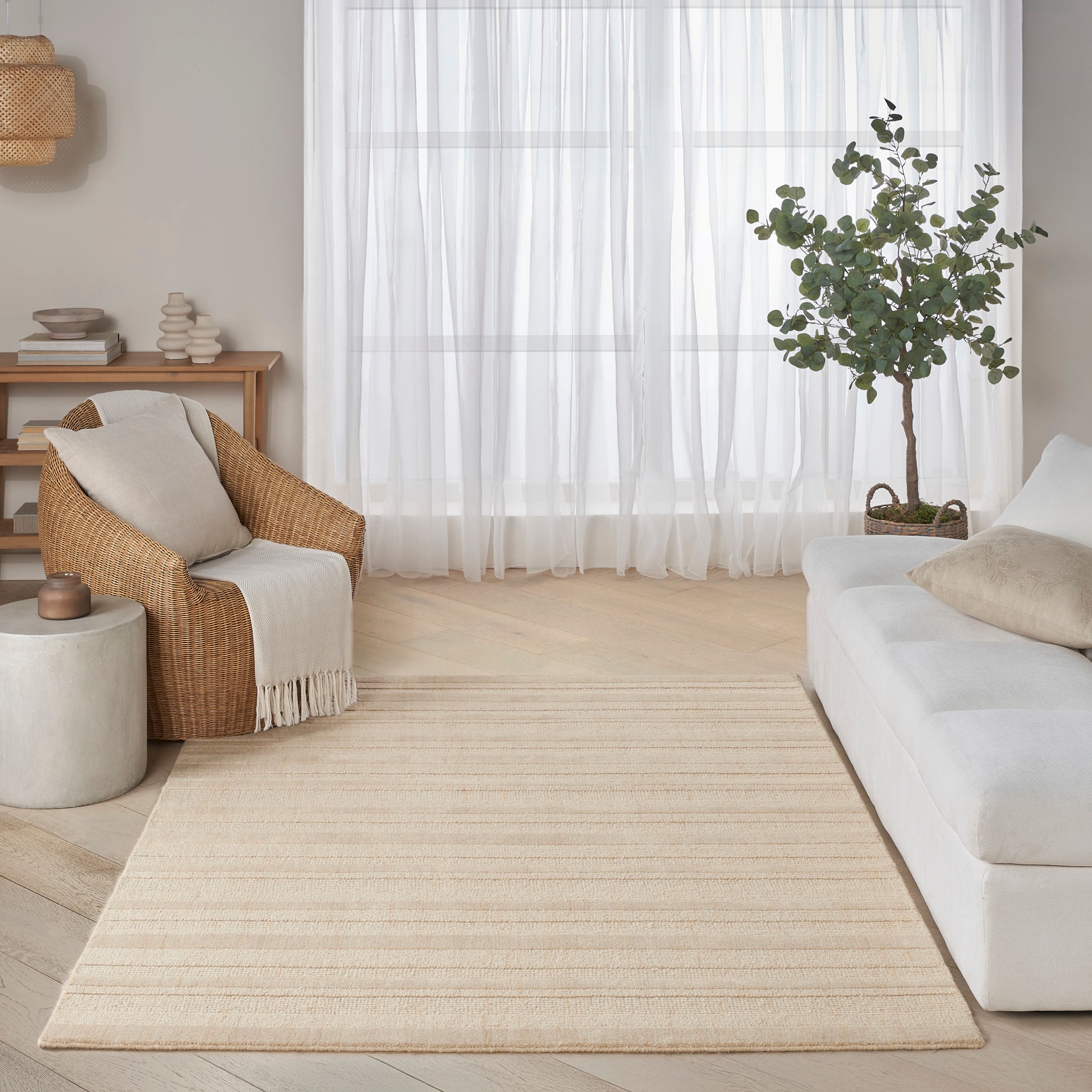 Bright neutral living room with beige woven rug, light wood floor, white sofa, wicker armchair, sheer curtains, potted tree.