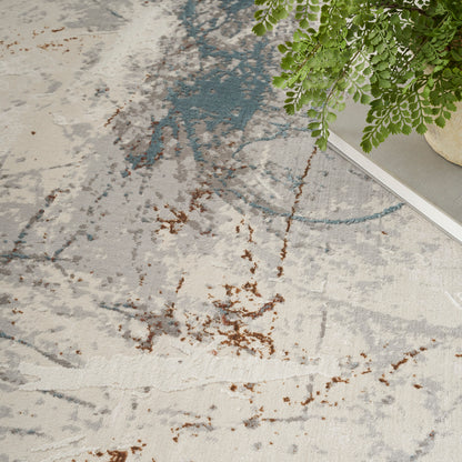 Soft abstract area rug in cream with gray and teal arcs and rust-brown specks beside a green plant.