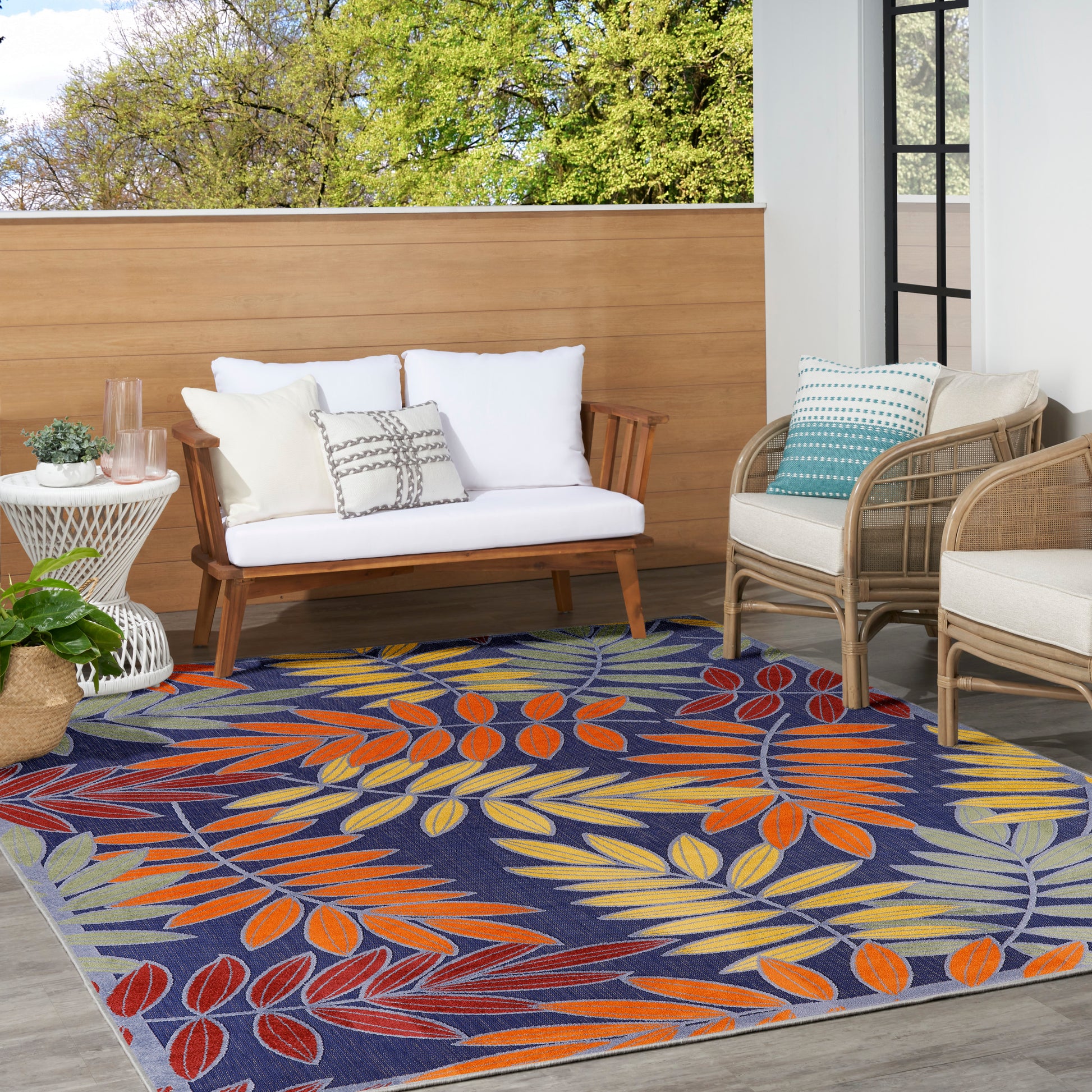 Outdoor patio with navy rug featuring orange, yellow and teal leaf pattern; white cushioned wood sofa and wicker chairs.