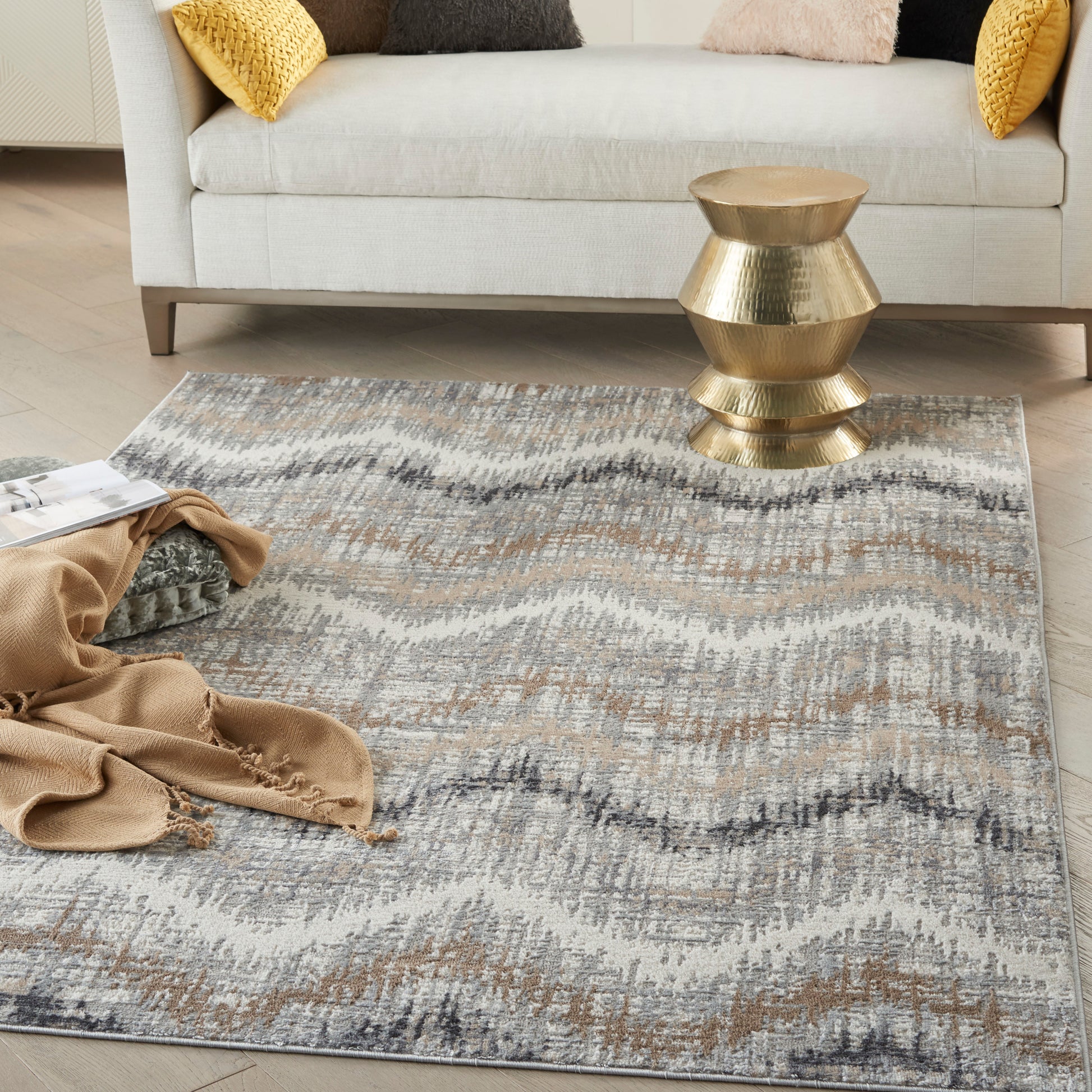 Gray-beige wavy patterned area rug in a bright living room with a gold geometric side table, cream sofa, and mustard-yellow cushions.