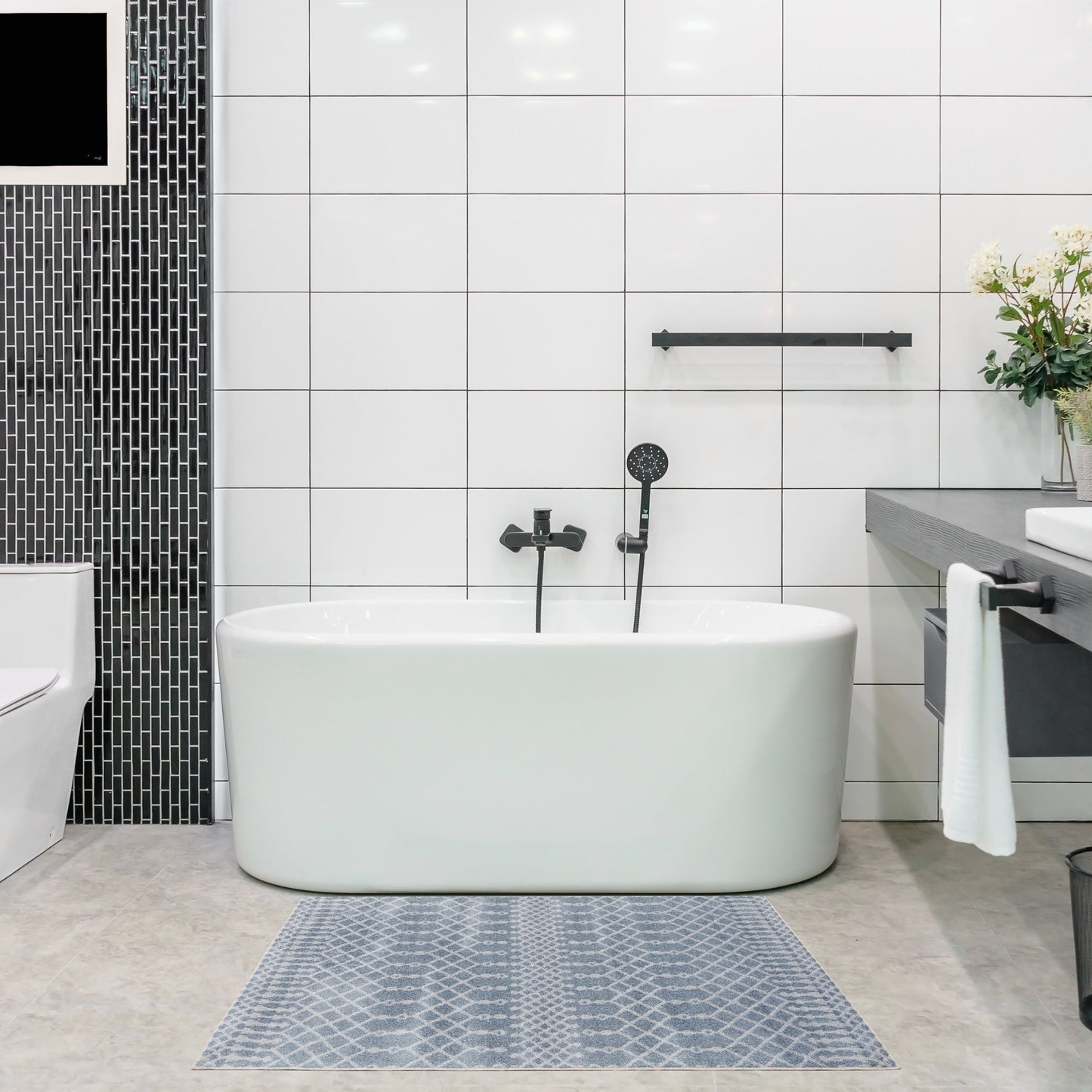 White freestanding oval bathtub with matte black fixtures in a modern white-tiled bathroom; blue-gray geometric rug.