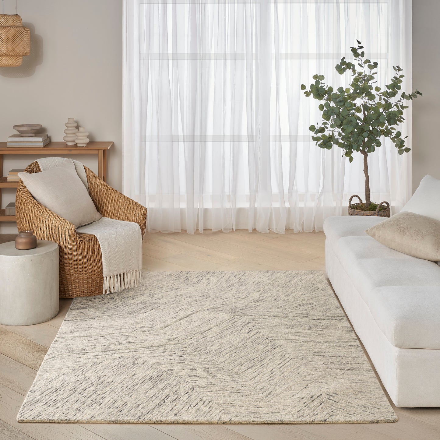 Cozy living room with cream sofa, woven armchair, beige textured rug, light wood floors, sheer curtains, potted tree.
