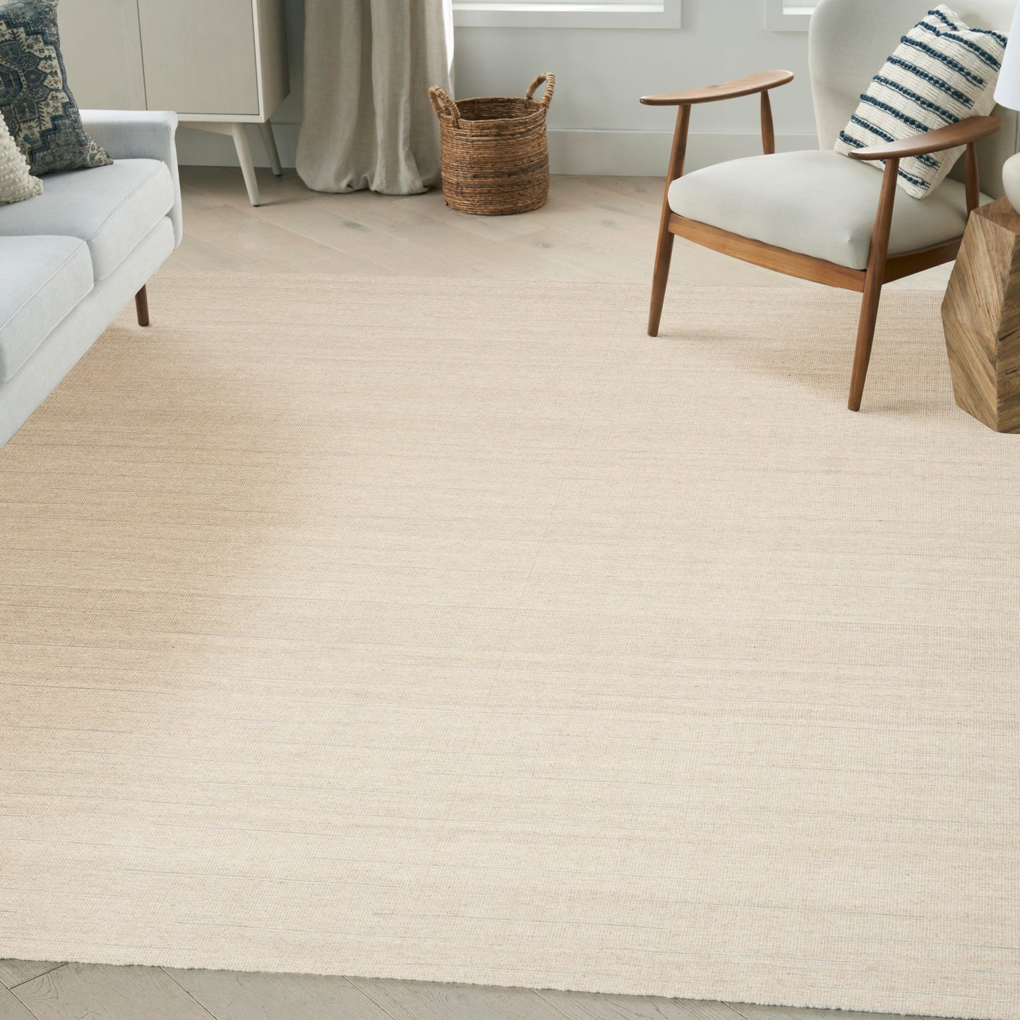 Bright living room with light beige area rug, mid-century wood chair, pale sofa, woven basket, & natural light.