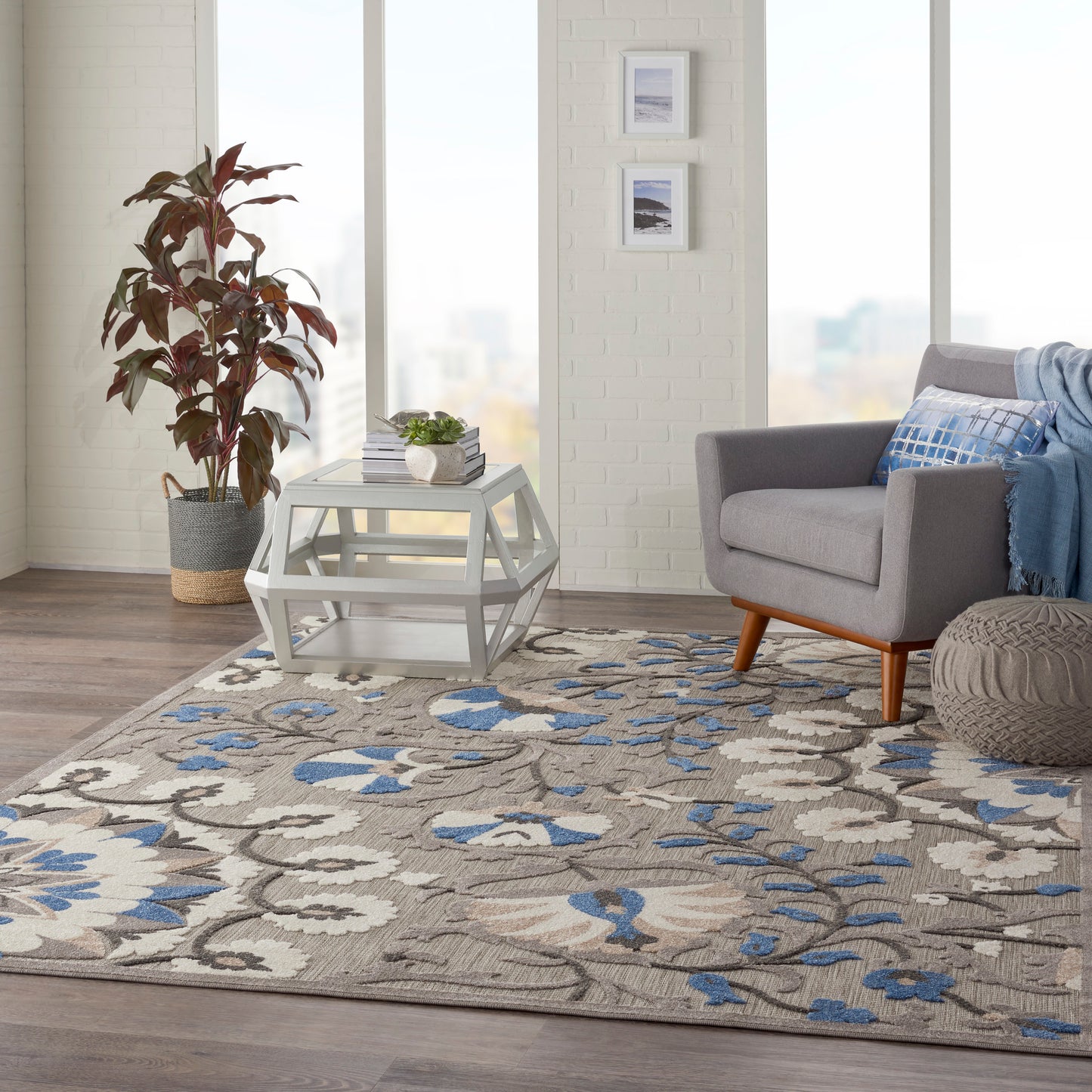 Bright living room with a beige-blue floral area rug, geometric white coffee table, gray sofa with blue throw, potted plant.