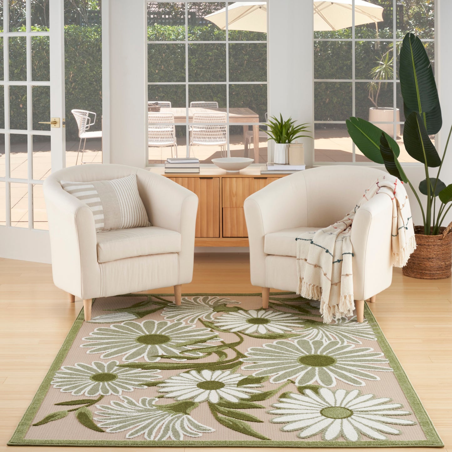 Two cream fabric armchairs with light wood legs sit on a beige rug with white daisies & green leaves in a bright living room.