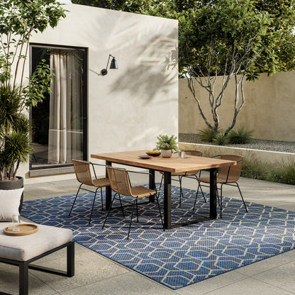 Outdoor dining set: wooden table with black metal legs and wicker chairs on a blue geometric rug, beige walls and greenery.