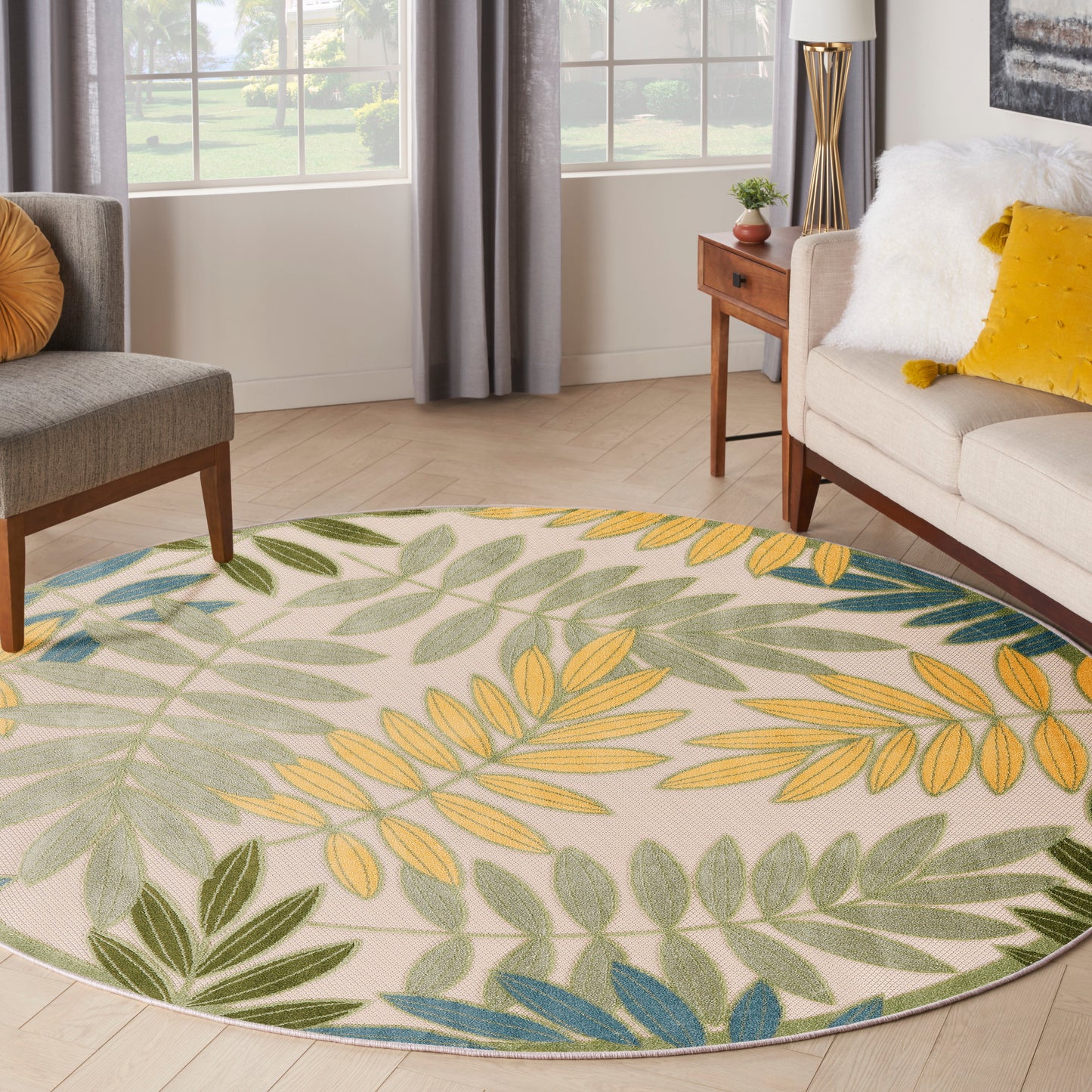 Round botanical rug with yellow, green and blue leaves on beige background; living room setup with beige sofa and gray chair.