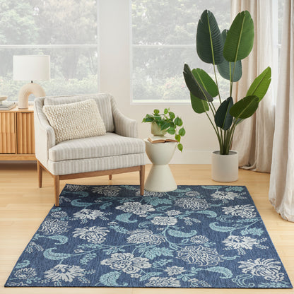 Blue floral-pattern area rug on light wood floor beside beige striped armchair, white side table, and large potted plant.