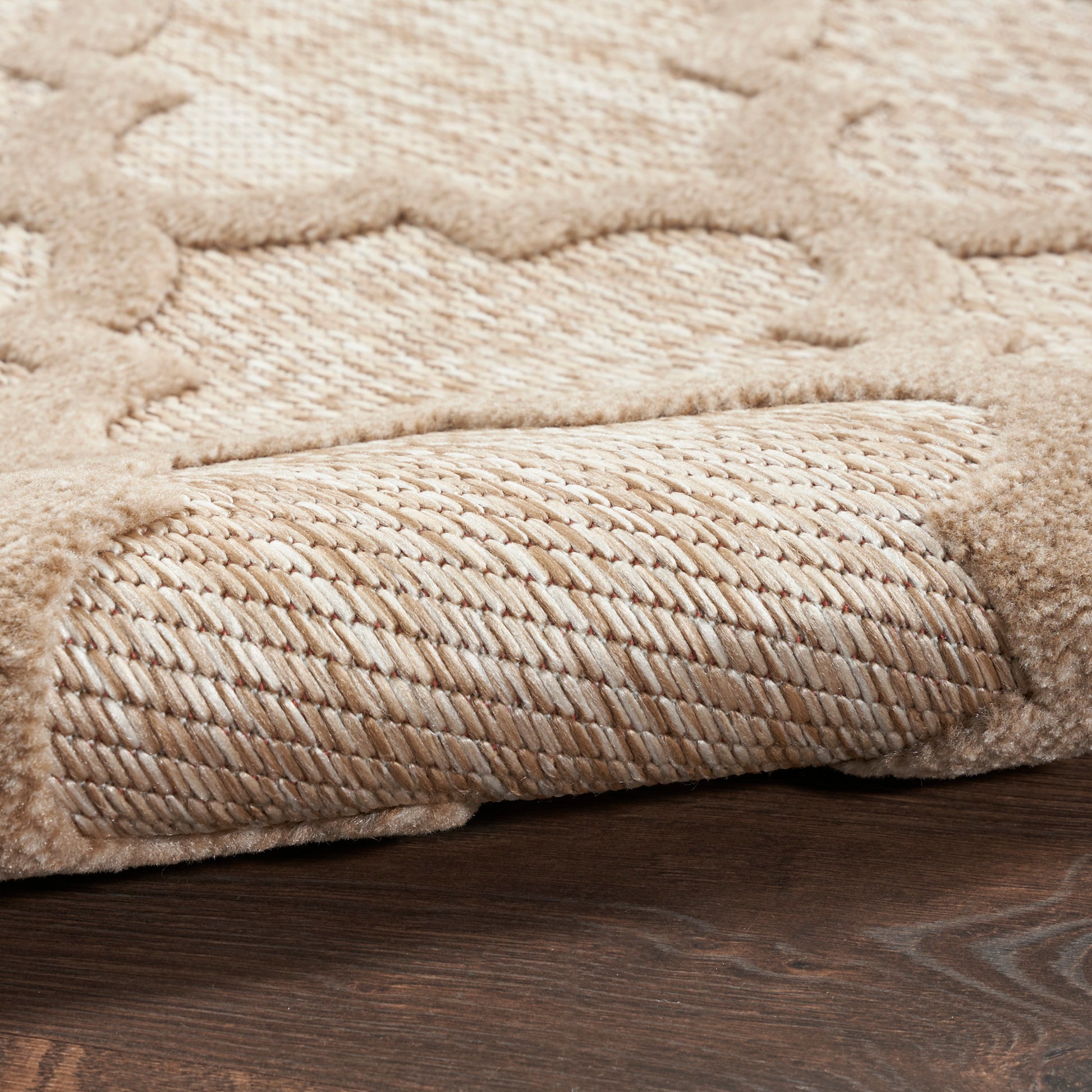 Beige plush shag area rug with decorative pattern; soft pile, rolled edge reveals braided underside on dark wood floor.