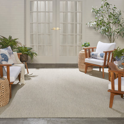 Bright sunroom with white cushioned wooden armchairs, blue patterned pillows, wicker tables, plants, and a beige woven rug.