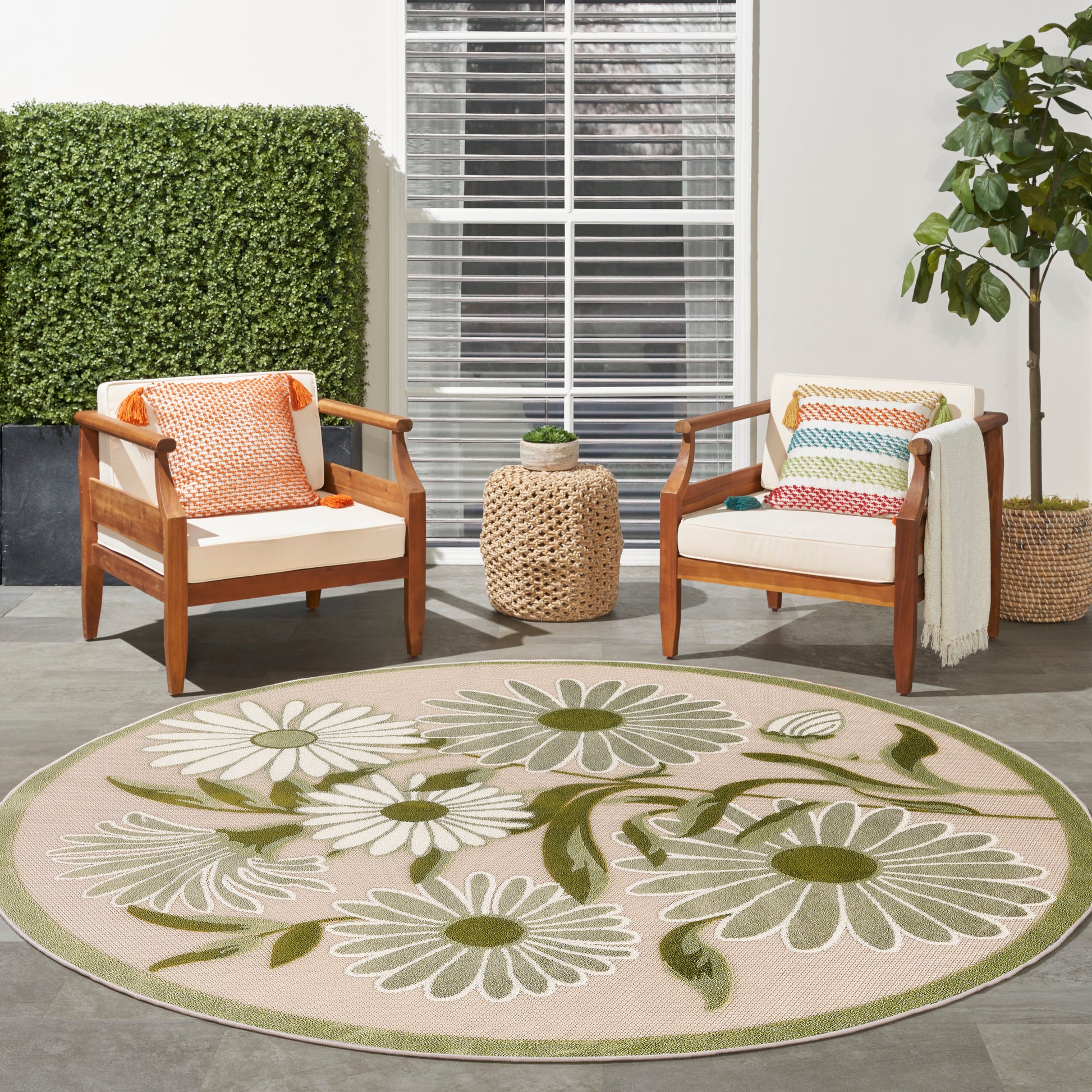 Outdoor patio set: round daisy rug, two wooden armchairs with beige cushions, colorful pillows, woven side table.