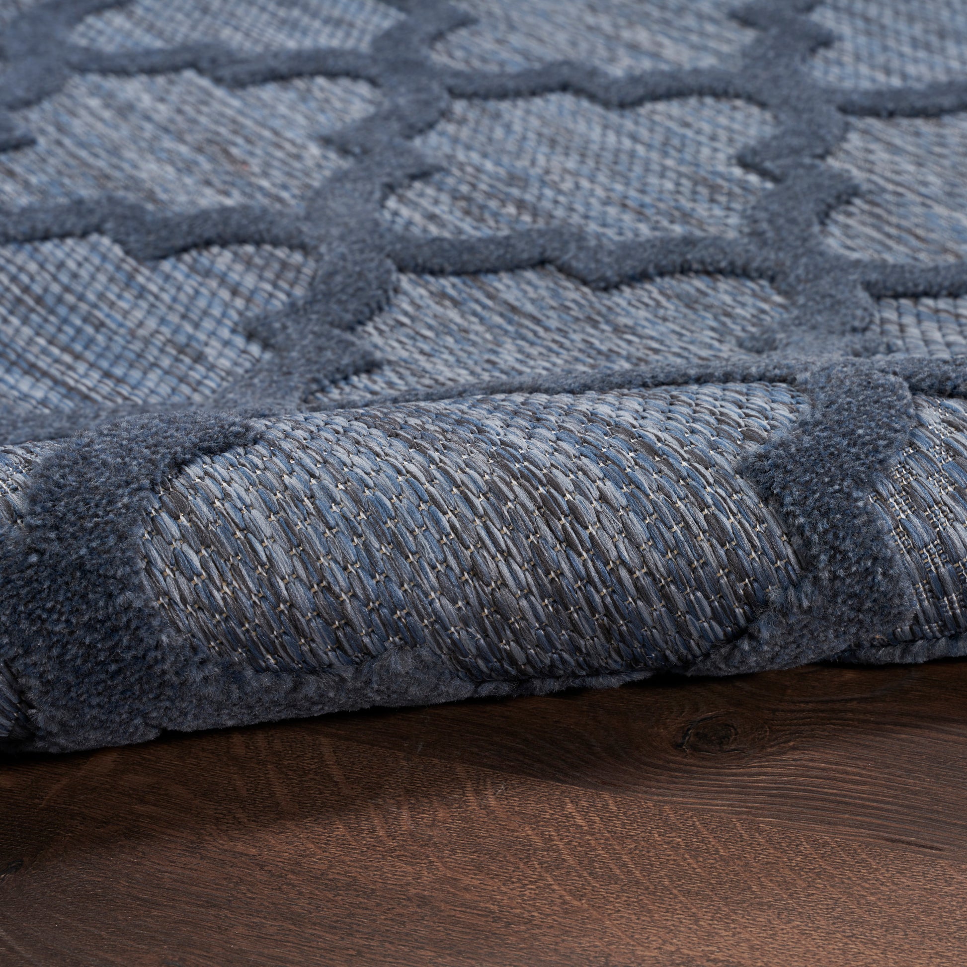 Blue-gray rug with geometric lattice pattern and soft woven pile, rolled edge on wood floor.