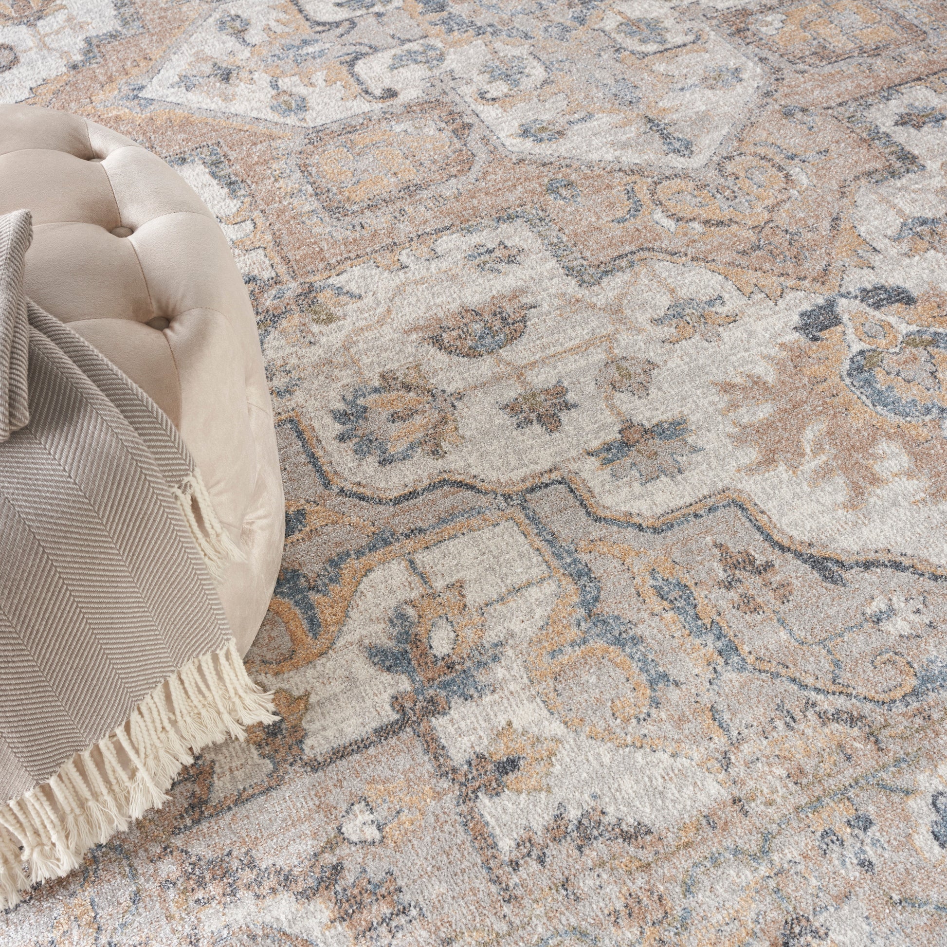 Beige round tufted ottoman with striped fringed throw draped over it on a patterned beige-blue rug.