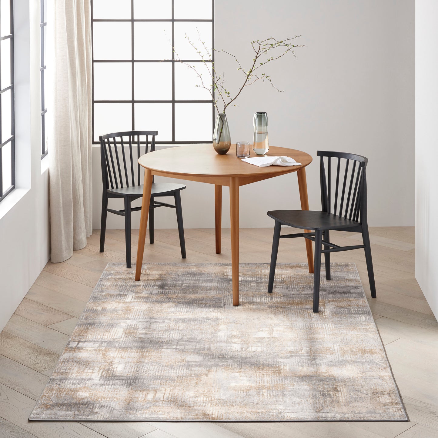 Beige-gray rug on floor with a glass carafe and a vase with branches; partial view of a round dining table and two black spindle chairs.