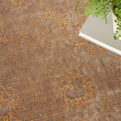 Warm-toned oriental rug with brown, gold and taupe floral vines; top-right shows a light gray notebook and green fern.