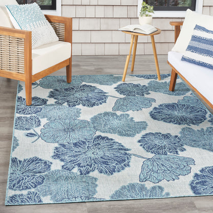 Blue floral area rug with large leaf shapes on light background; wood-framed chairs with white cushions and a round table.