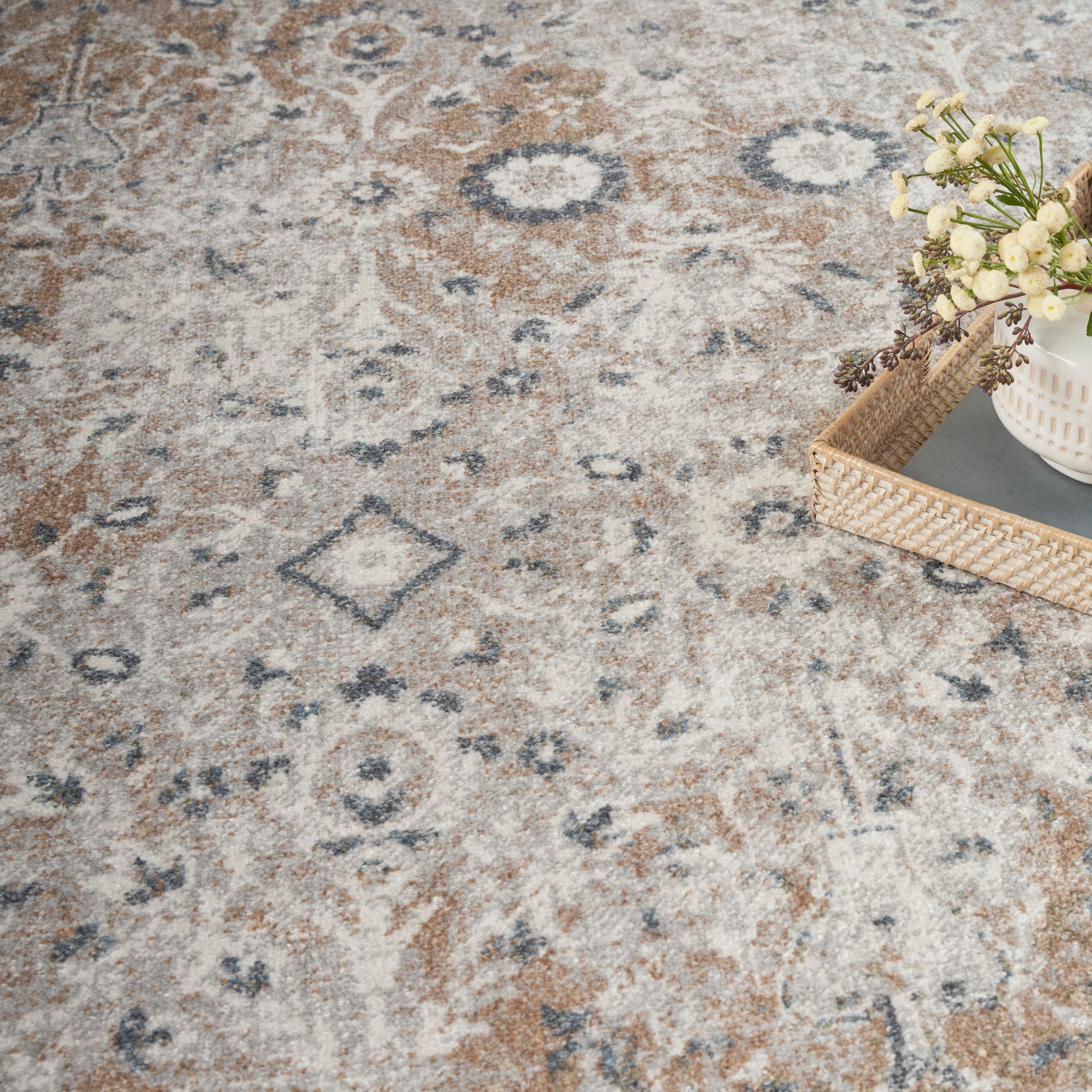 Neutral patterned area rug in beige, gray, blue and rust tones; woven tray with a white vase and cream flowers.
