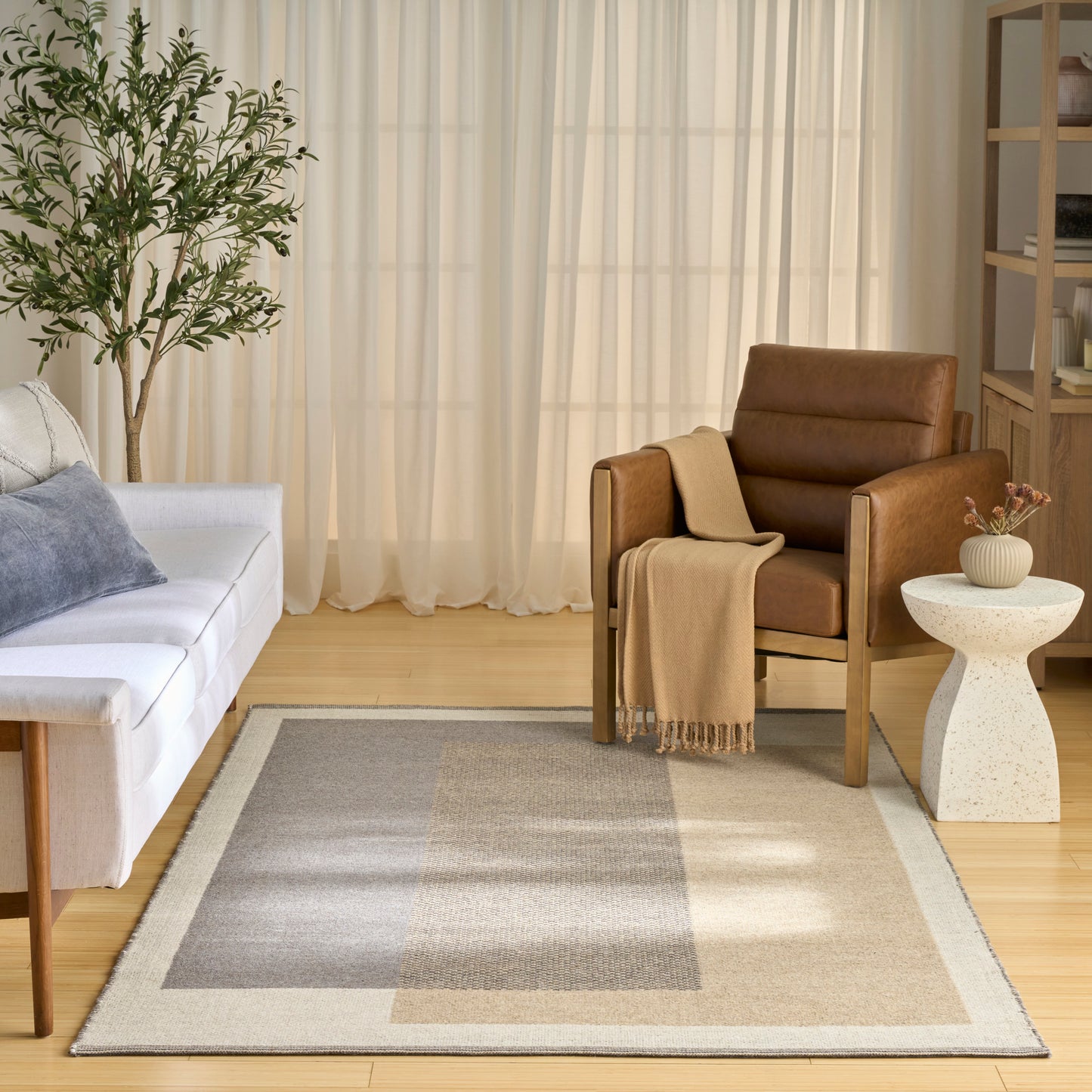 Cozy living room: white sofa, gray cushions, brown leather armchair with beige throw, beige-gray rug, plant, sheer curtains.
