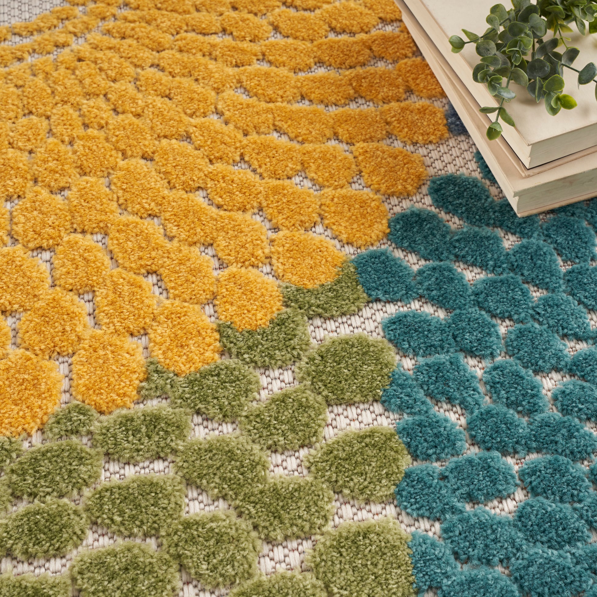 Colorful tufted rectangular rug with mustard yellow, olive green, and teal blue pom-poms on a woven base; books and a plant nearby.