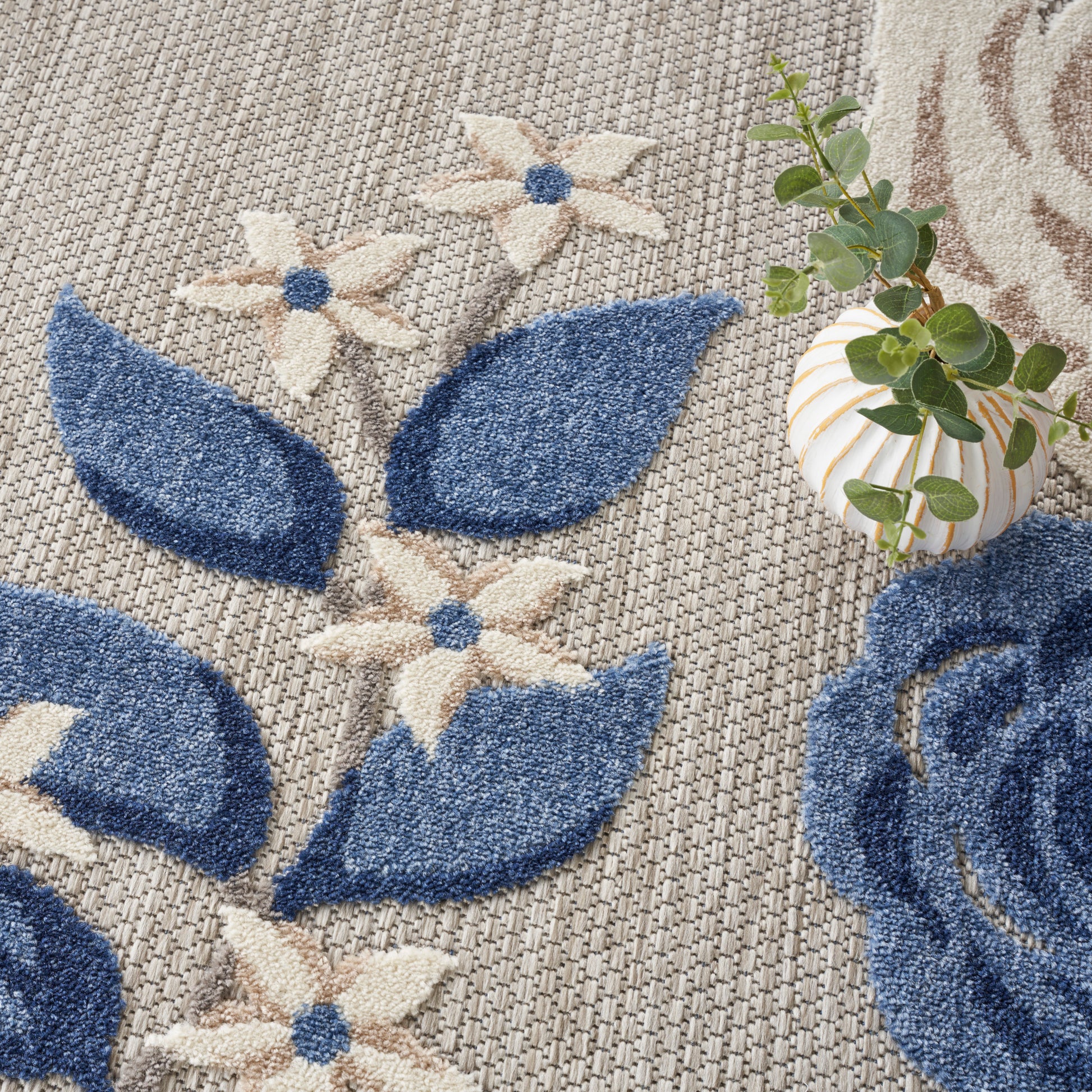 Blue and cream floral woven rug with large blue petals and cream flowers with blue centers on beige textured background.