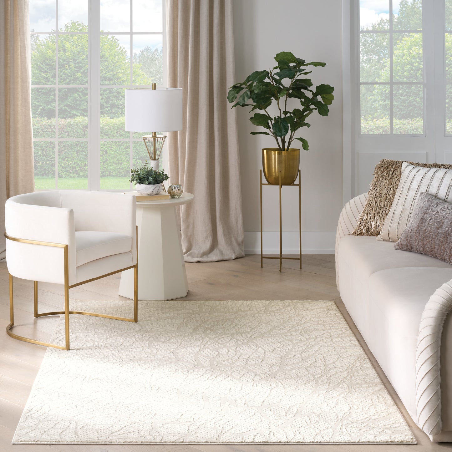 Neutral living room with white sofa and gold-frame chair, round side table, plant, cream rug and large sunny windows.