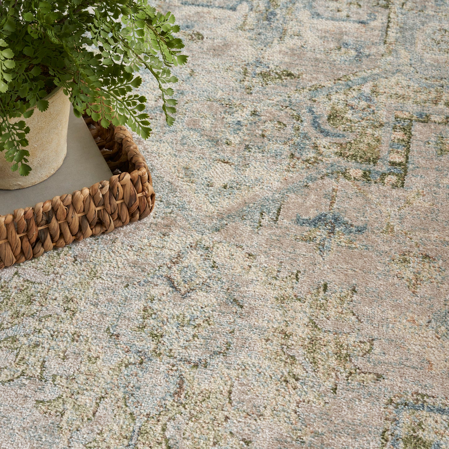 Fern in beige pot on a woven basket atop a pastel blue-pink-green patterned rug.