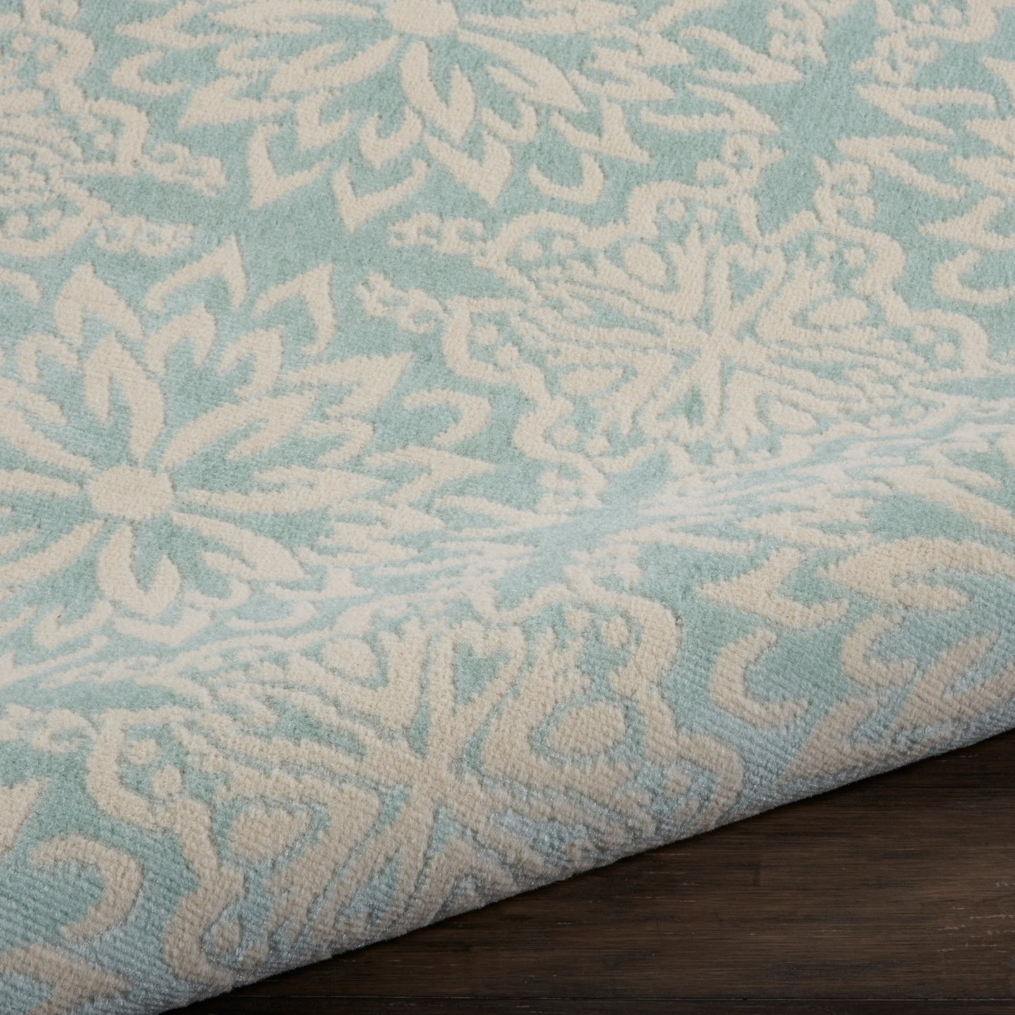 Turquoise and beige damask-patterned rug sample with a folded edge on a dark wood table.