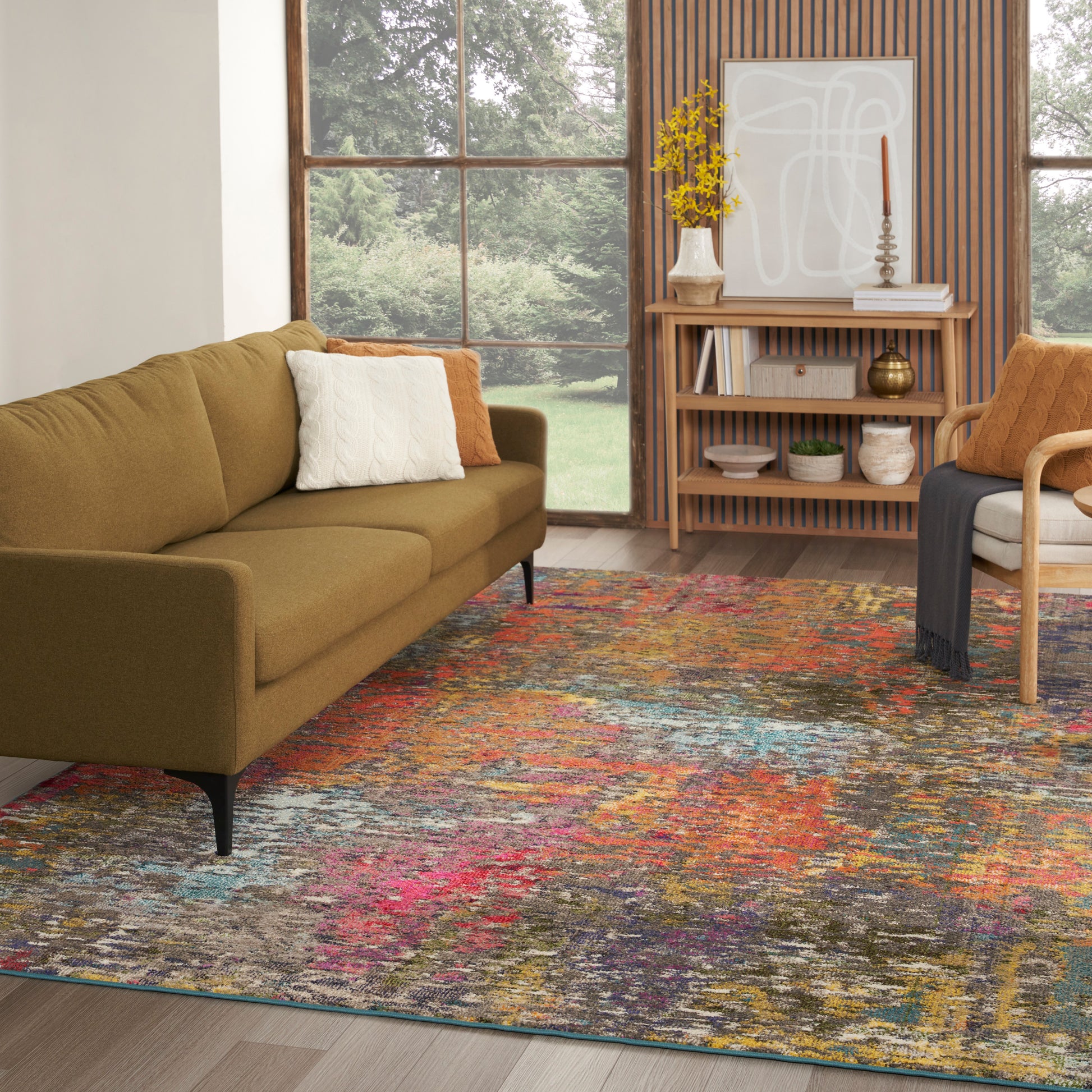 Olive-green mid-century sofa with white and orange pillows, colorful abstract rug, wooden shelves in bright living room.