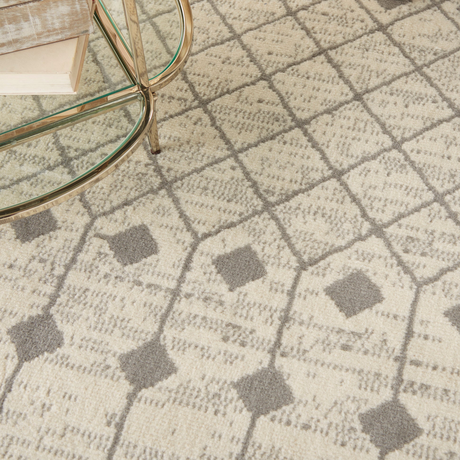 Beige patterned area rug with gray diamond motif under a glass coffee table with brass legs and a stack of books