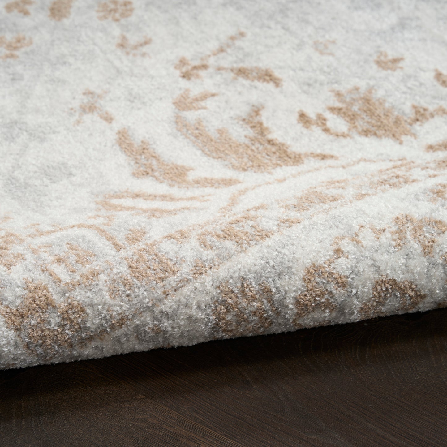 Cream and beige damask-patterned rug with plush pile, curled edge on dark wooden floor.