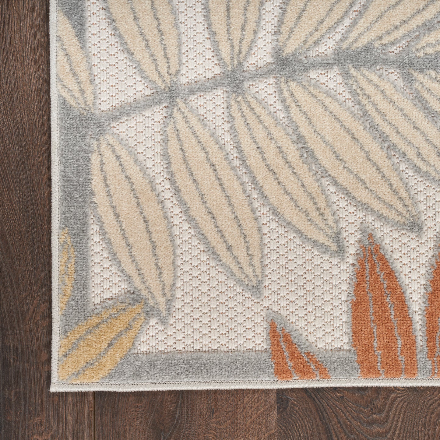 Close-up of a textured woven rug corner with beige and gray leaf motifs, orange accents, on dark wood floor.