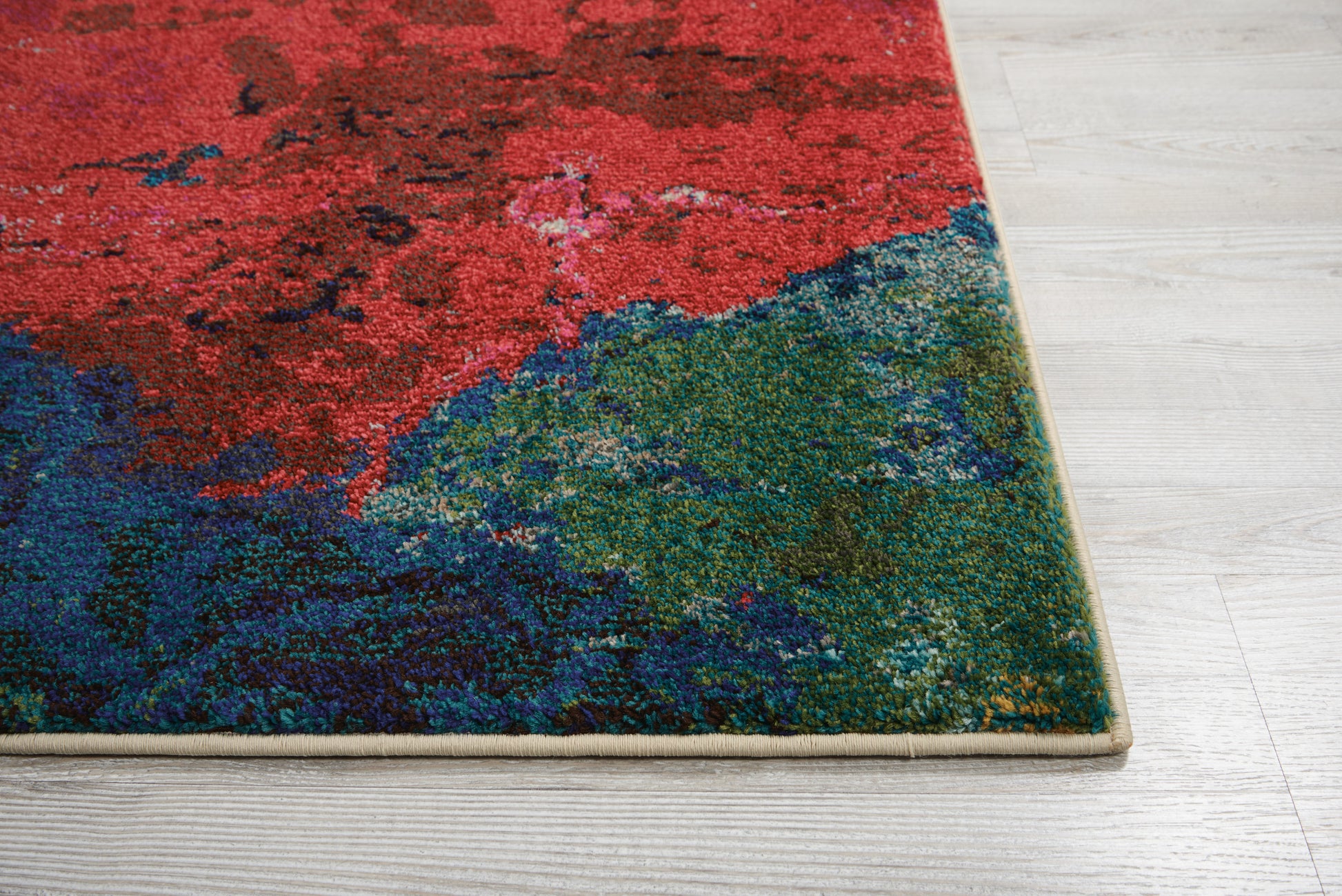 Abstract patterned area rug with red, blue and green hues and a beige edge on light wood flooring.