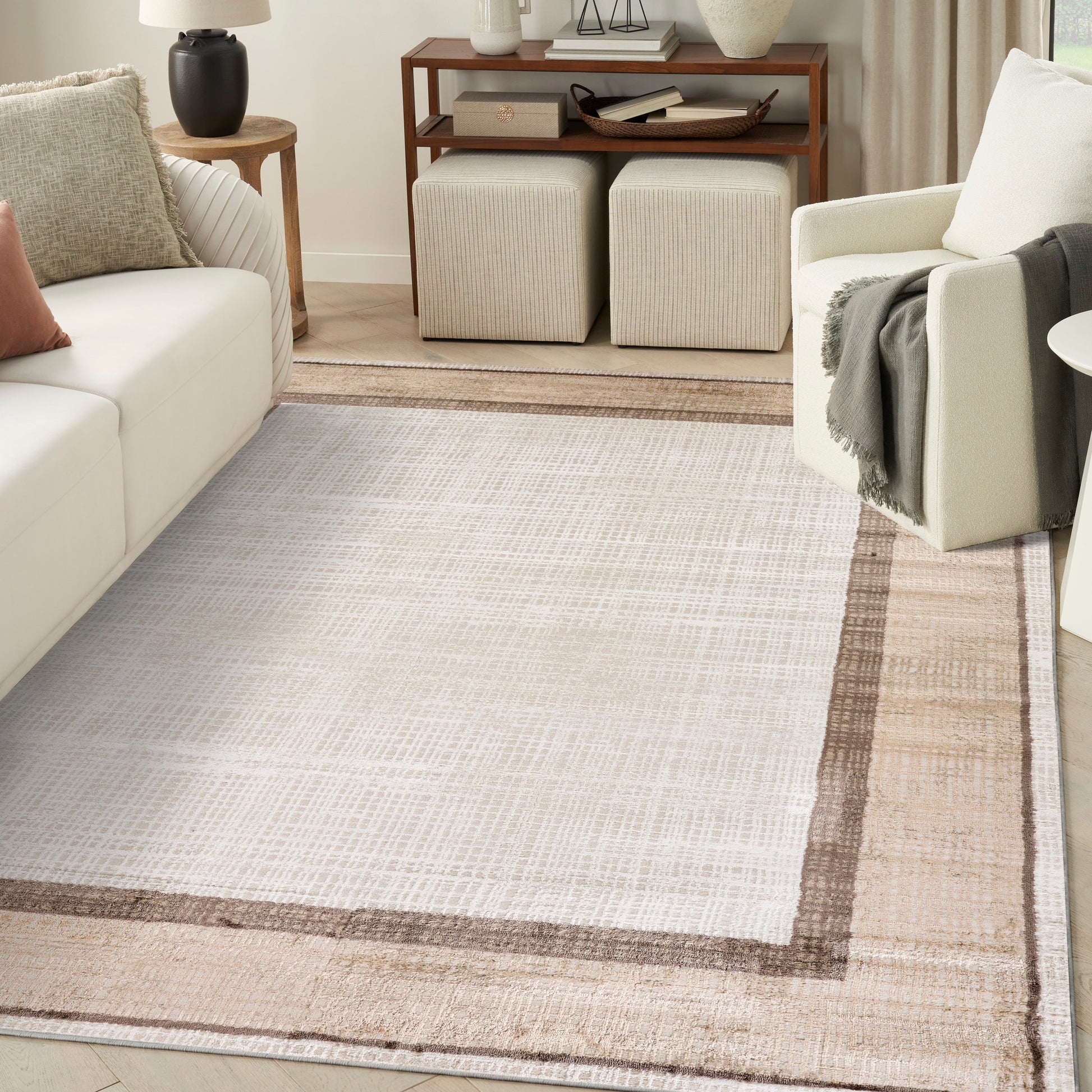 Neutral woven area rug in light beige with a tan border, paired with a cream sofa, armchair, and warm wood console.